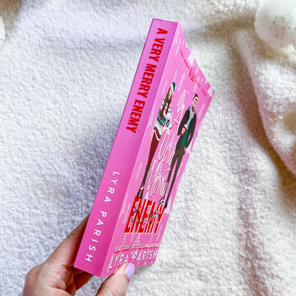 A hand holds a pink copy of the book 'A Very Merry Enemy' by Lyra Parish, featuring illustrations of characters on the cover, set against a soft white textured blanket background.