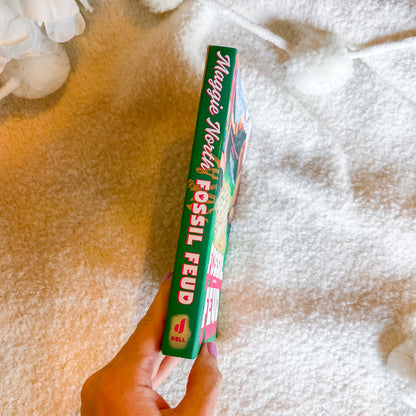 A hand holding a book titled 'Fossil Feud' against a soft white textured blanket background.
