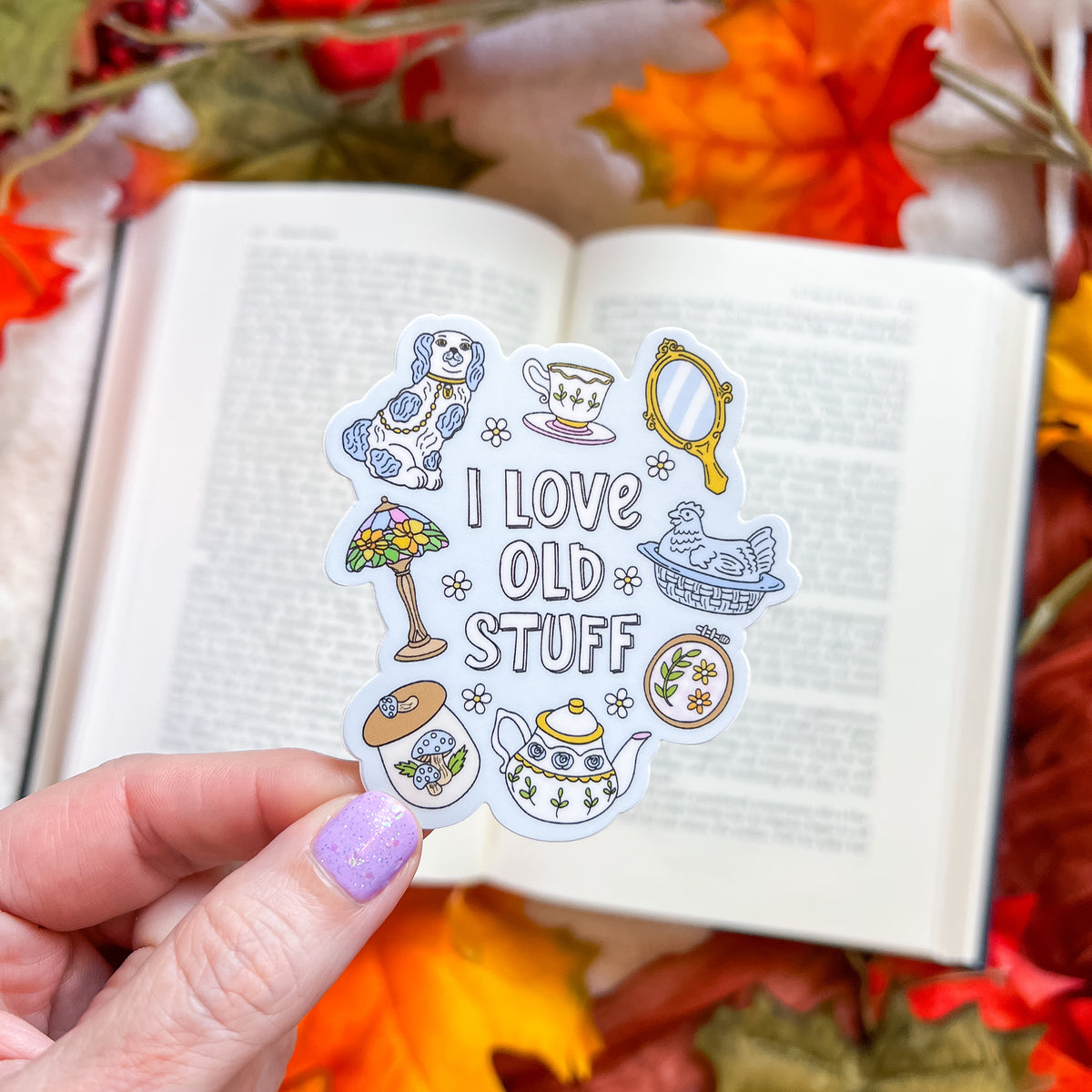 A hand with lavender nail polish holds a die-cut sticker featuring a vintage tea-time theme and the text 'I LOVE OLD STUFF'. The sticker includes illustrations of a dog, a teacup, a mirror, a lamp, a cat, a teapot, and a small box.