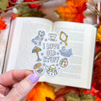 A hand with lavender nail polish holds a die-cut sticker featuring a vintage tea-time theme and the text 'I LOVE OLD STUFF'. The sticker includes illustrations of a dog, a teacup, a mirror, a lamp, a cat, a teapot, and a small box.