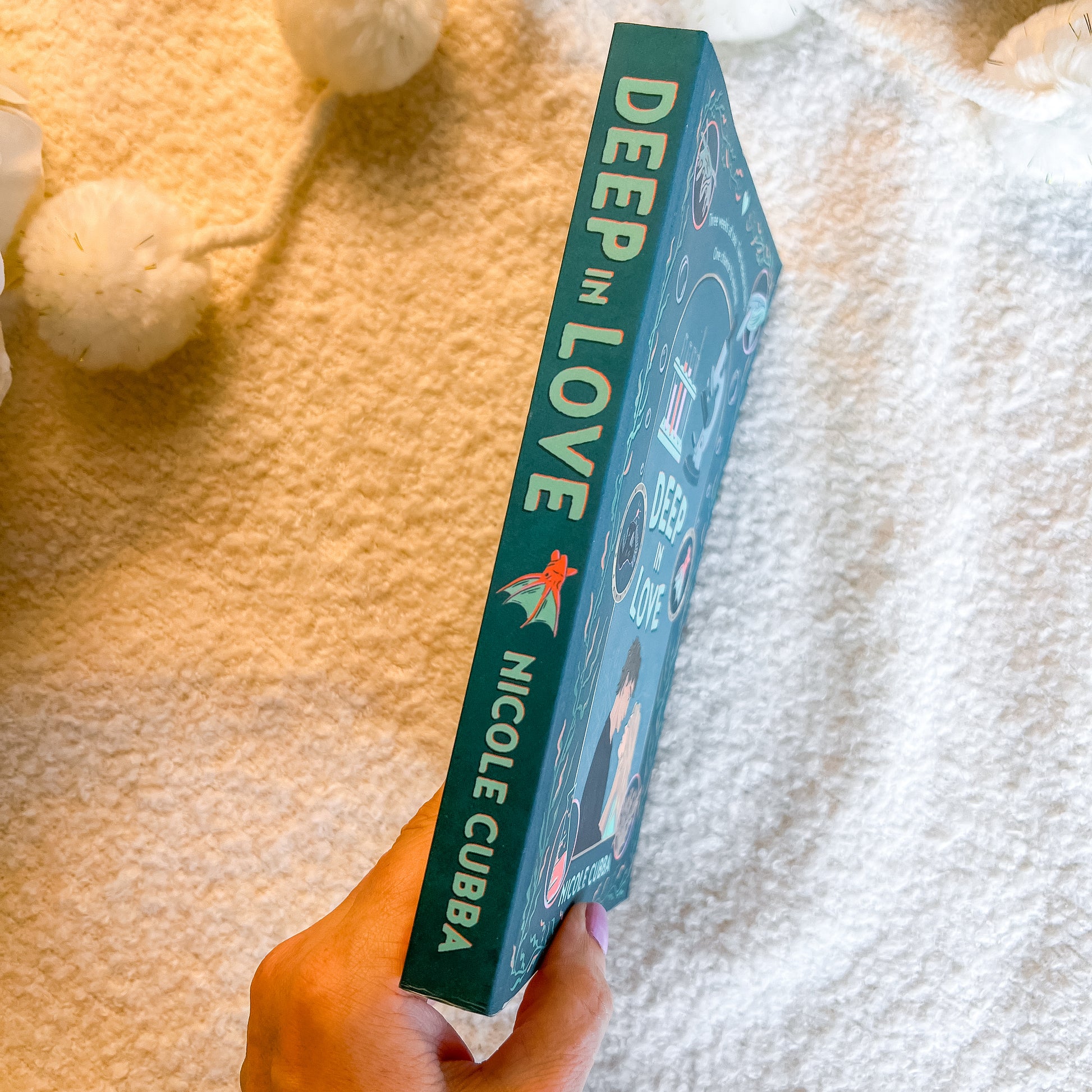 A hand holds a teal book titled 'DEEP IN LOVE' by Nicole Cuba, standing vertically against a soft white textured blanket background with decorative white pom-poms.
