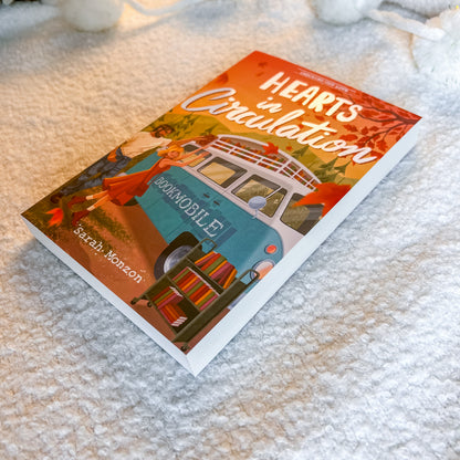 A book titled "Hearts in Circulation" by Sarah Monson lies on a soft white textured blanket. The cover features an illustration of a teal and white camper van with the word "BEDMOBILE" on the side, surrounded by autumn leaves and a woman in a red dress.