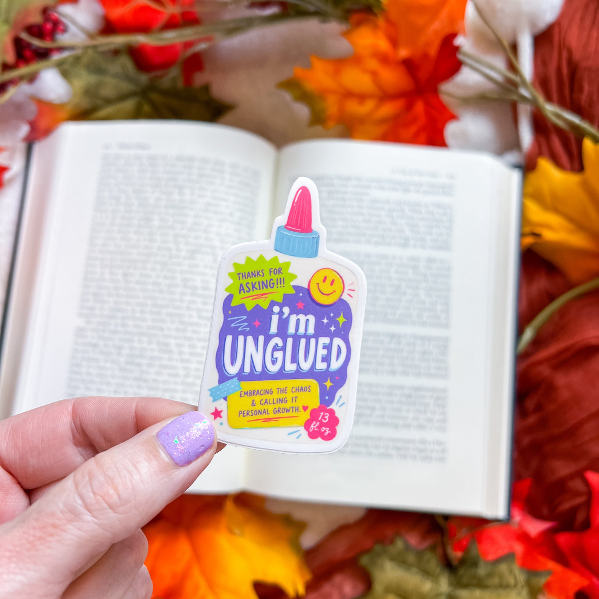 A hand with lavender glitter nail polish holds a die-cut sticker shaped like a glue bottle in front of an open book, surrounded by autumn leaves. The sticker features the text 'i'm UNGLUED' with a purple background, yellow smiley face, and pink glue cap.