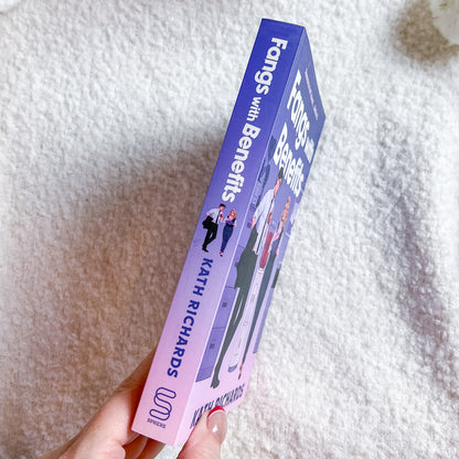 A hand holds a book titled "Fangs with Benefits" by Kath Richards. The book cover features a purple-to-pink gradient with illustrations of people and bold white text, set against a soft white textured blanket background.