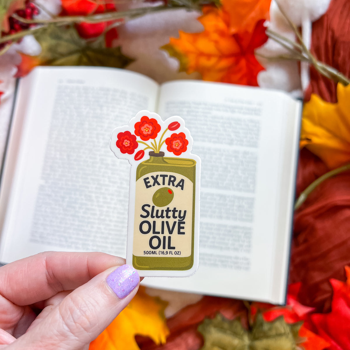 A hand with lavender glitter nail polish holds a die-cut sticker shaped like an olive oil bottle. The sticker reads 'EXTRA Slutty OLIVE OIL' with red flowers on top.