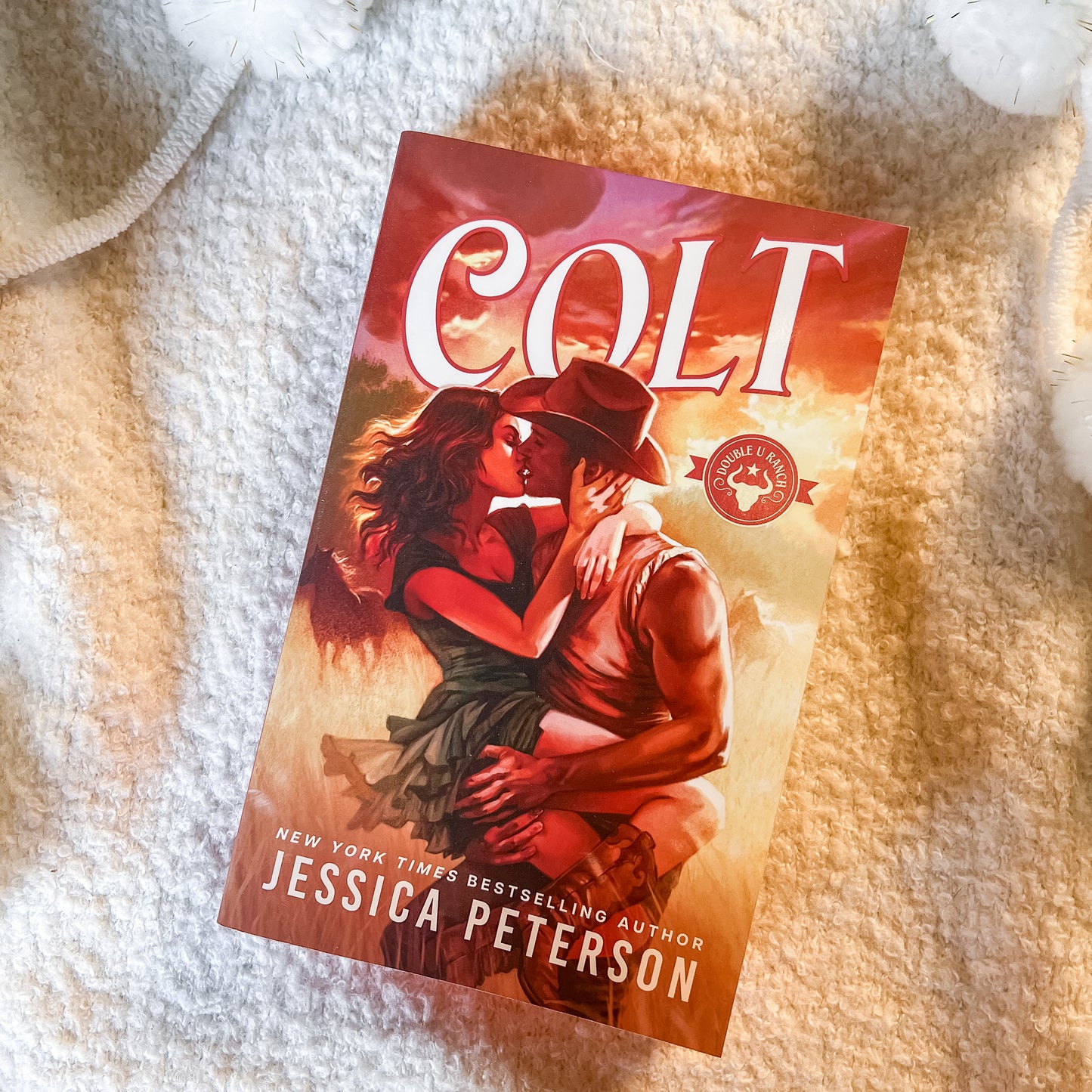A book titled 'COLT' by Jessica Peterson lies diagonally on a soft, textured cream-colored blanket. The cover features a romantic illustration of a couple embracing under a warm sunset sky, with the title 'COLT' in large white letters at the top and the author's name at the bottom.