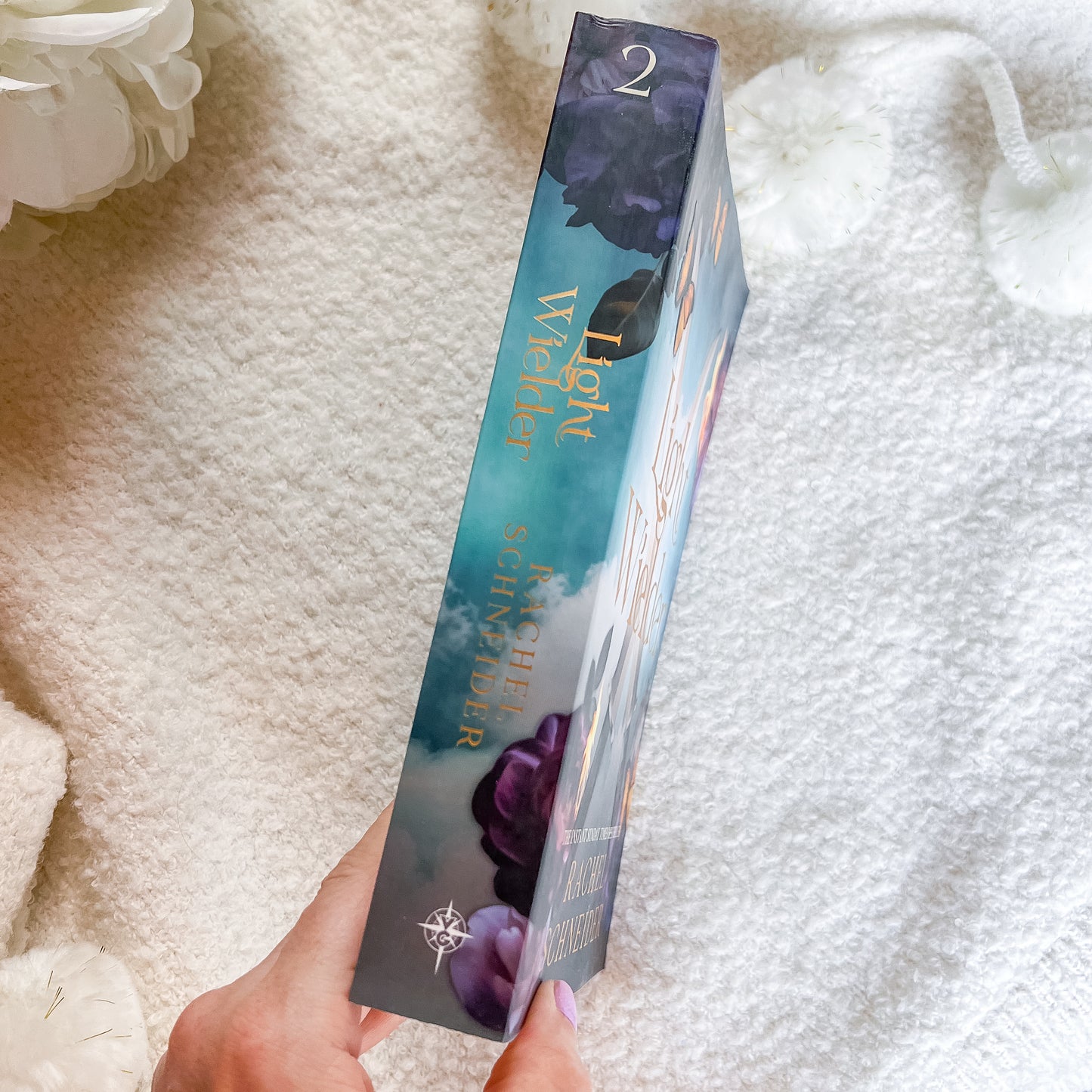 A hand holds a book showing the spine with purple rose illustrations and gold lettering, set against a soft white textured blanket 