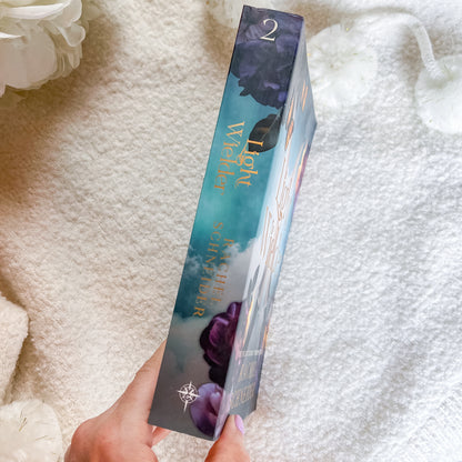 A hand holds a book showing the spine with purple rose illustrations and gold lettering, set against a soft white textured blanket 