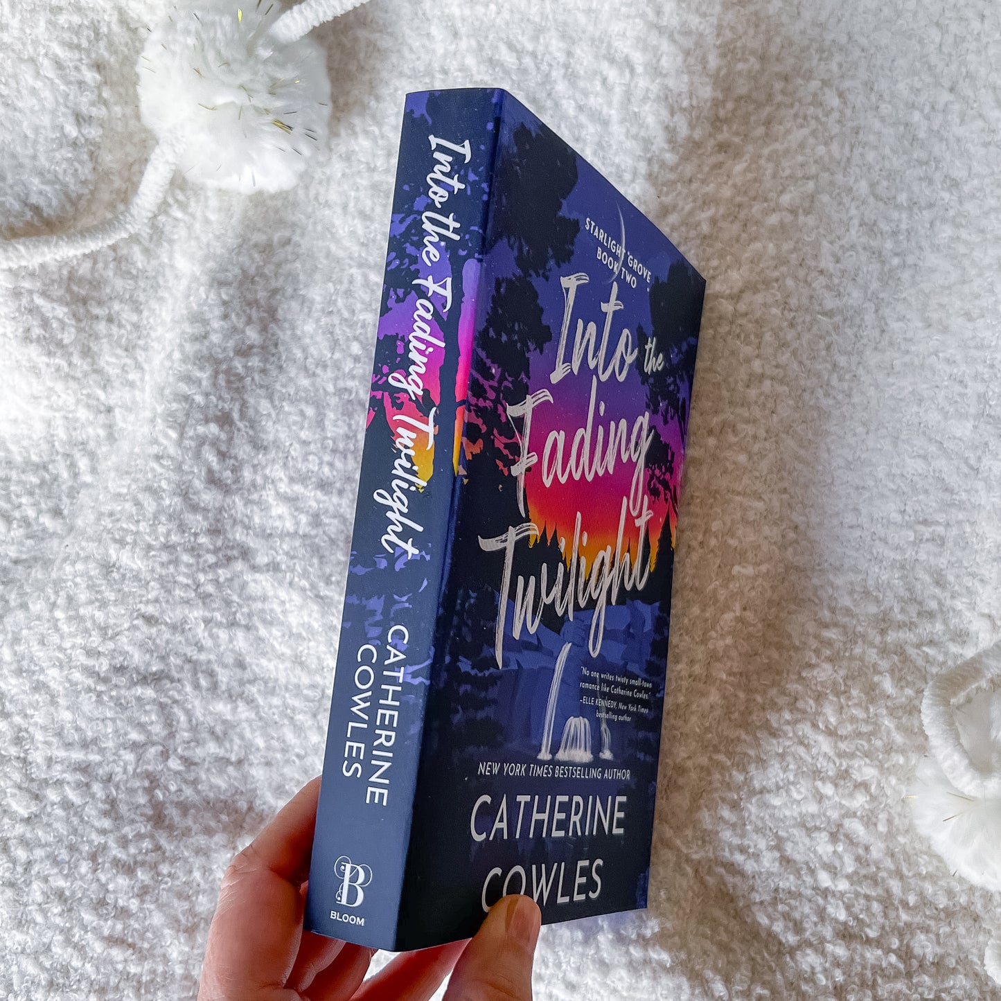 A hand holds a copy of the book 'Into the Falling Twilight' by Catherine Cowles. The book cover features a dark blue and purple background with white title text and a colorful sunset gradient.