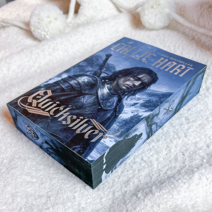A boxed book or card deck titled "Quicksilver" by Callie Hart, featuring artwork of a dark-haired armored figure against a snowy blue-toned background, photographed on a soft white textured blanket.
