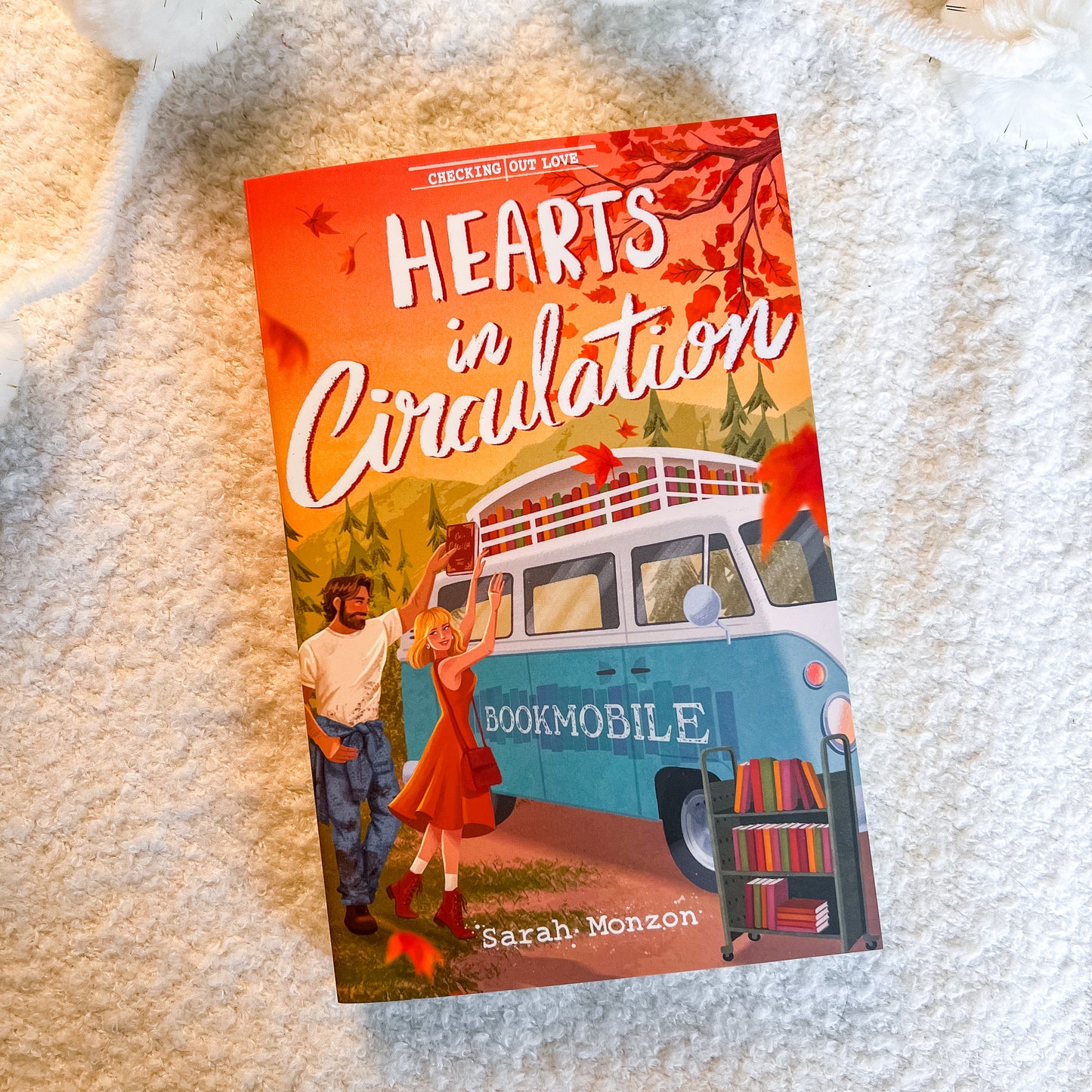 The book cover of 'Hearts in Circulation' by Sarah Monzon is displayed flat on a soft, textured white blanket. The cover art features a retro light blue bookmobile van, a couple standing joyfully, and red autumn leaves against a warm orange and yellow background.