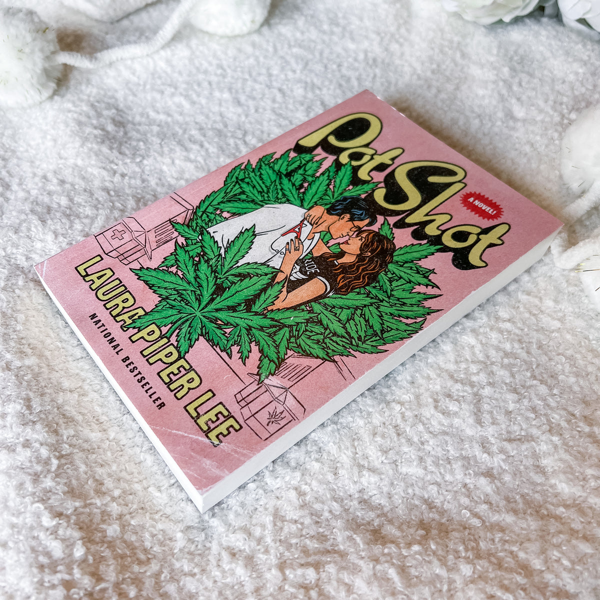 A  book titled 'Pot Shot' by Lauryn Piper Lee lies diagonally on a soft, textured white blanket. The cover features a pink background with a cartoon illustration of a woman surrounded by green cannabis leaves, and bold yellow and red typography.