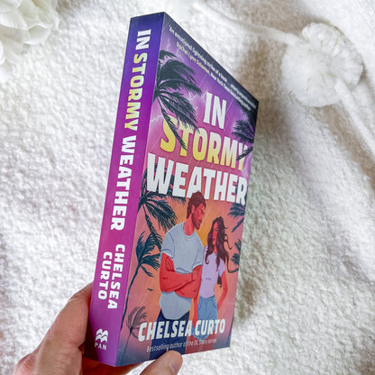 A hand holds a paperback book titled 'In Stormy Weather' by Chelsea Curto. The book cover features a purple-to-pink gradient with palm trees and an illustration of a couple, set against a soft white textured blanket background.