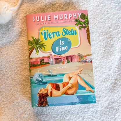 The book cover of 'Vera Stein Is Fine' by Julie Murphy, featuring a retro mid-century modern illustration of a woman in a pool with palm trees and a pink building, placed on a textured white blanket.