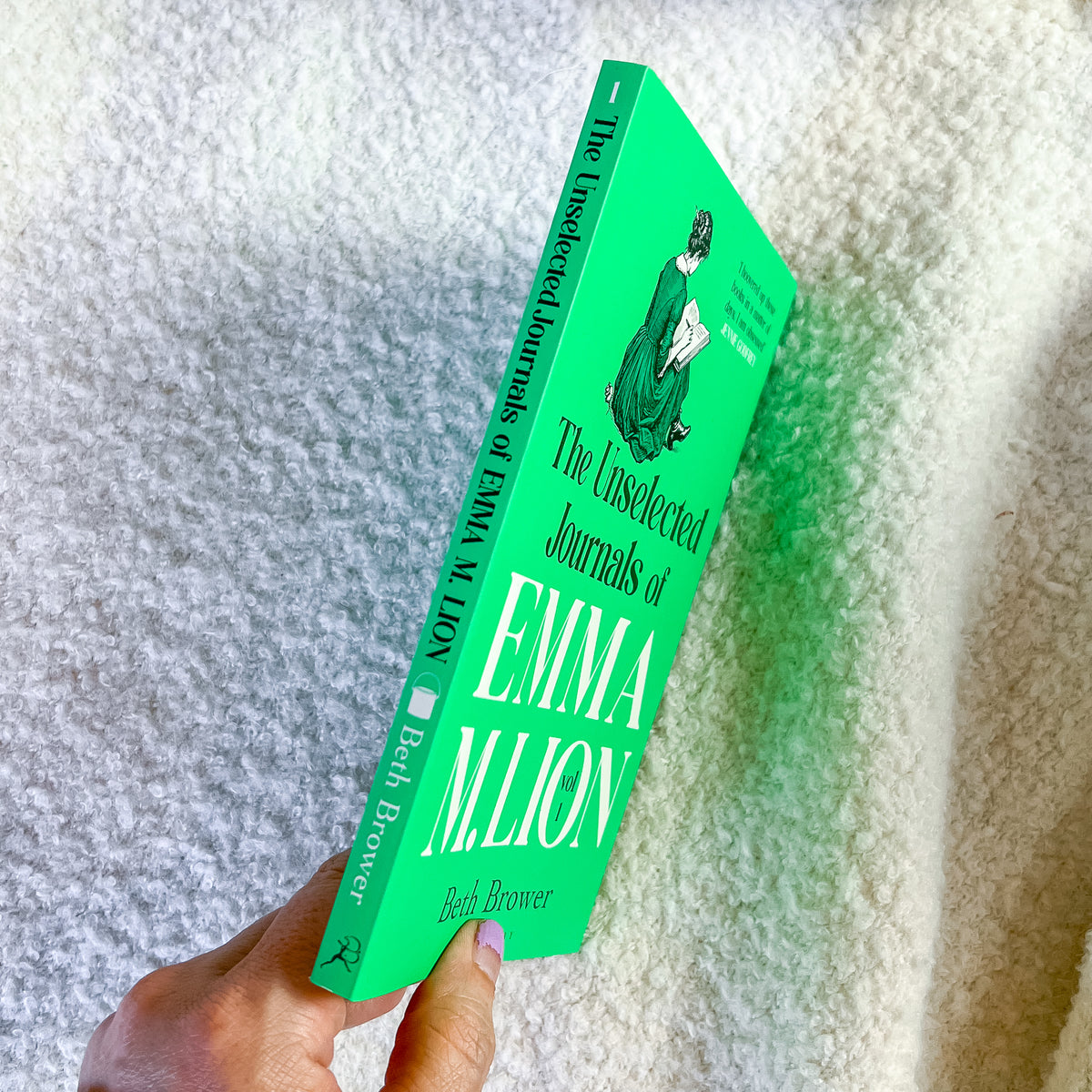 A hand holds a bright green hardcover book titled "The Unselected Journals of Emma M. Lion" by Beth Power, photographed against a textured off-white blanket.