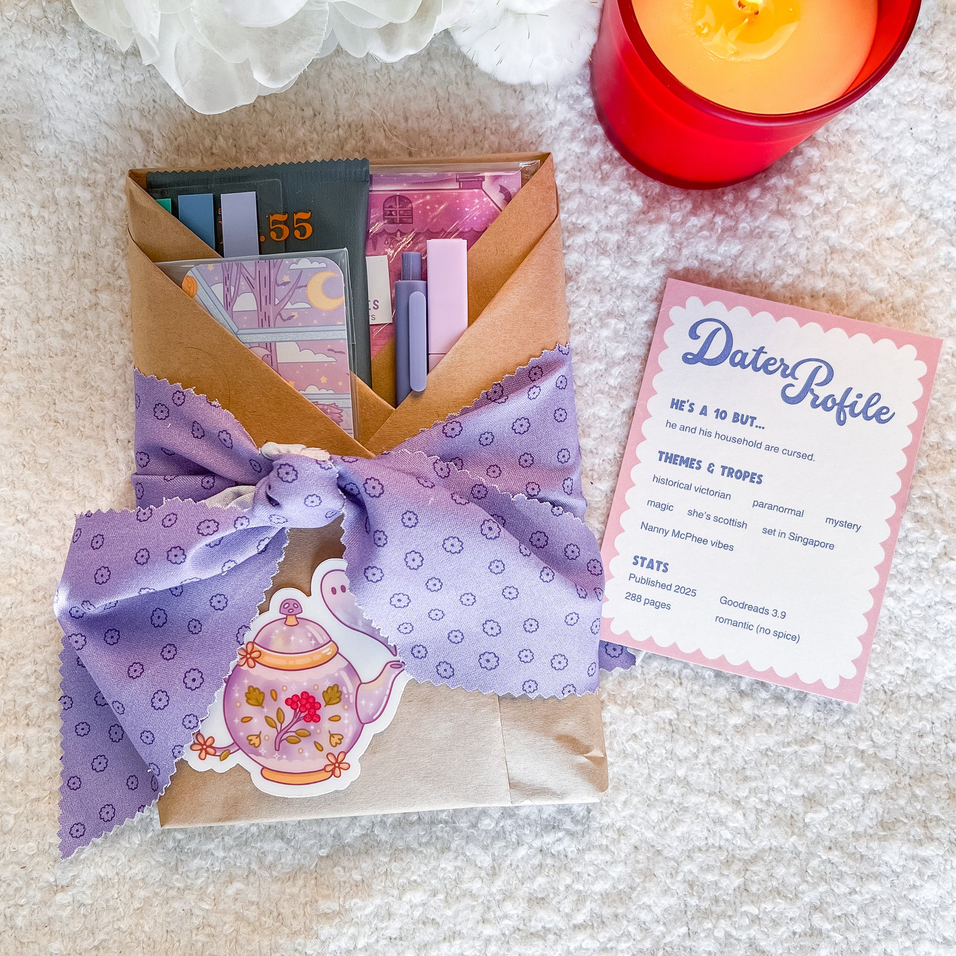 A styled flat-lay of a dater profile card and a wrapped book gift. The gift is wrapped in brown paper with a lavender patterned ribbon tied in a large bow, adorned with a floral teapot sticker.