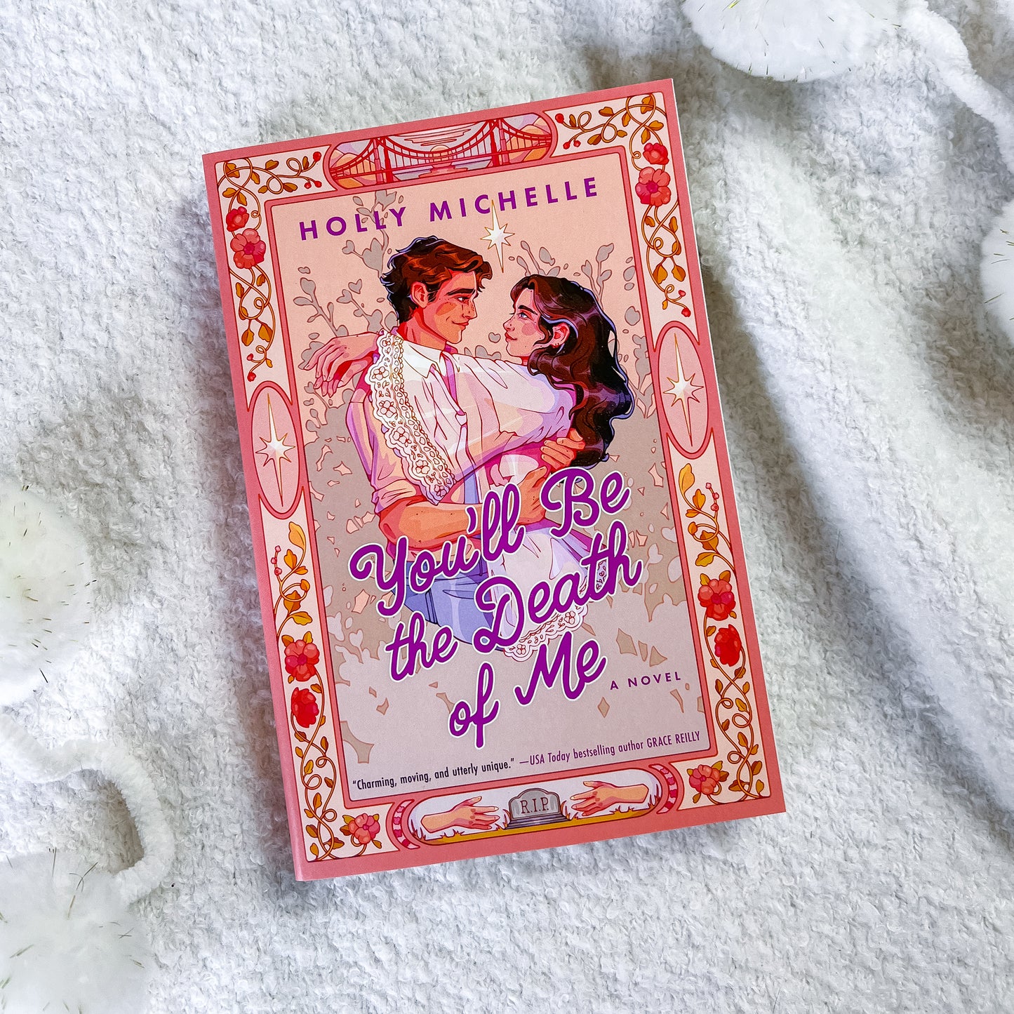 A book titled "You'll Be the Death of Me" by Holly Michelle, featuring a pink and red vintage-style cover illustration of a couple, placed on a white textured blanket.