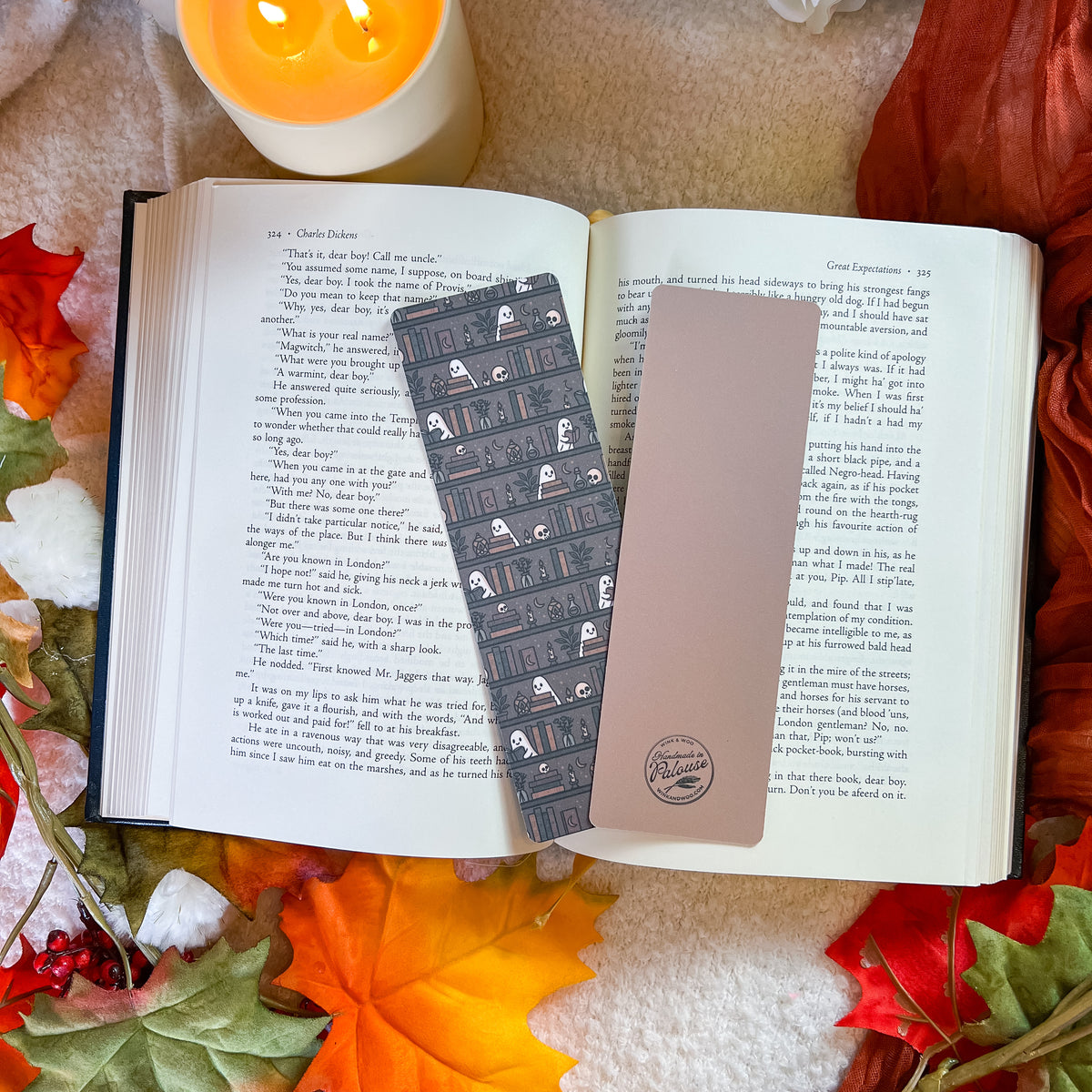 A decorative book bookmark with a dark gray and black pattern and white ghosts, alongside a solid light pink bookmark, displayed on open book pages surrounded by autumn leaves and a candle.