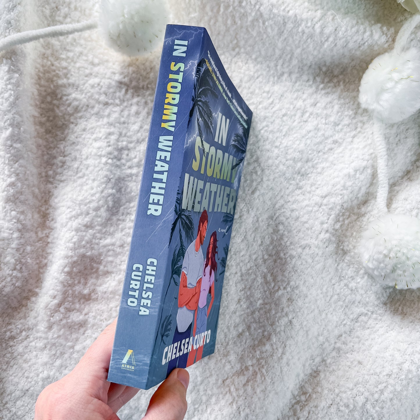 A hand holds a paperback book titled 'In Stormy Weather' by Chelsea Curto against a soft white textured blanket background.