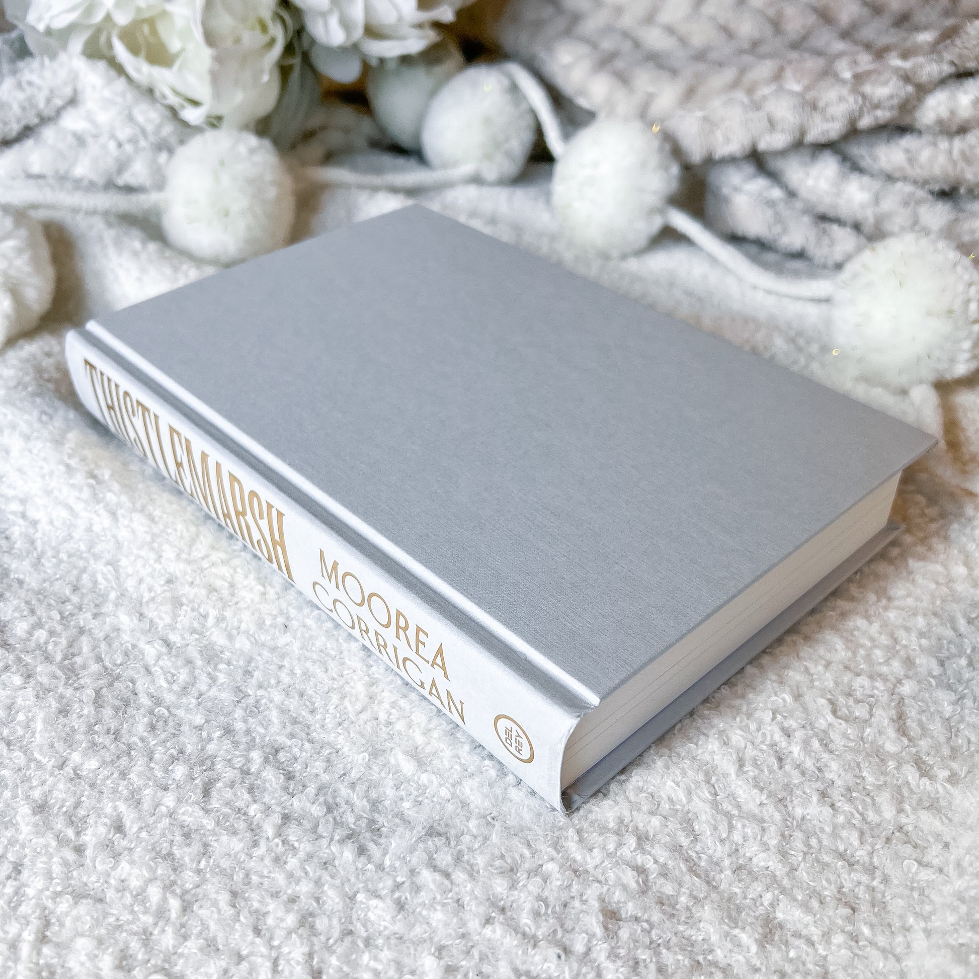 Gray book titled 'Thistlemarsh' by Moorea Morgan on a textured white surface with decorative elements.