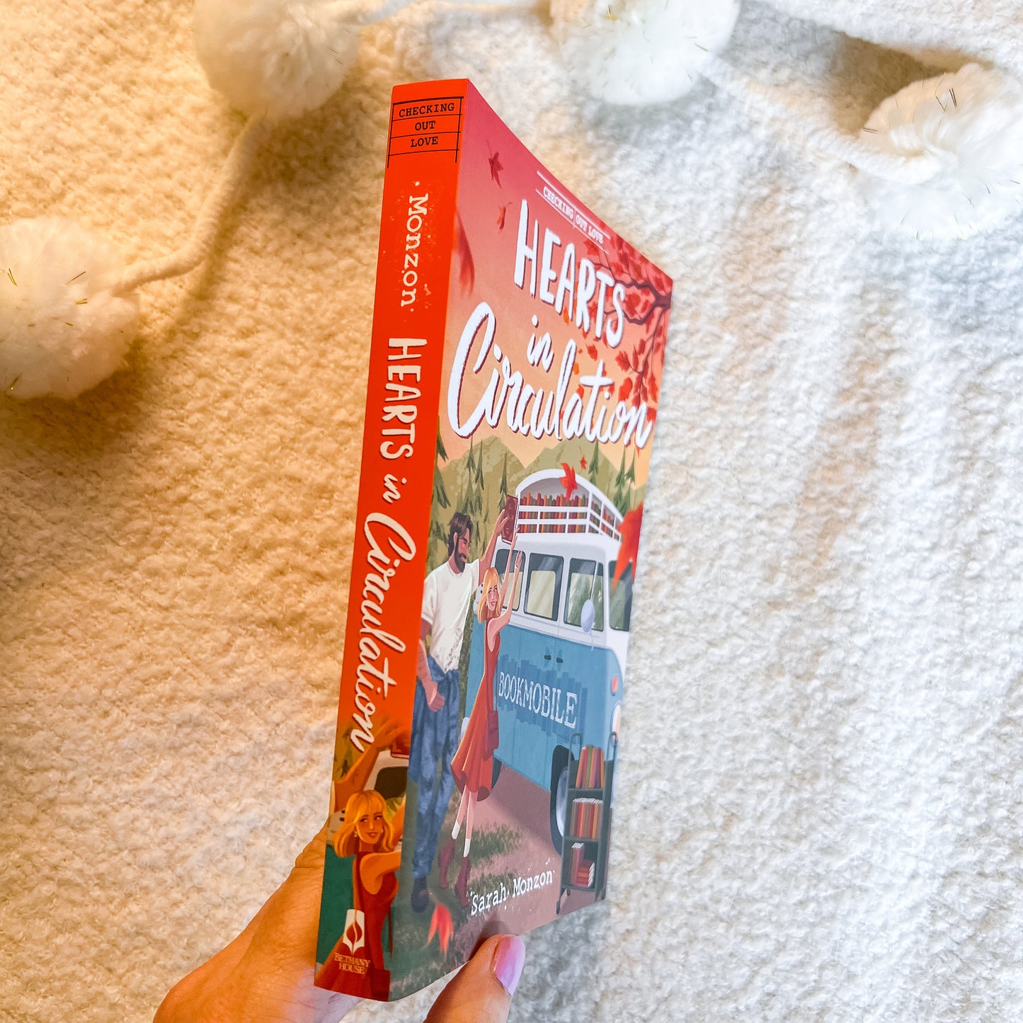 A hand holds a copy of the book 'Hearts in Circulation' by Sarah Dessen. The book cover features a vibrant red spine and cover with an illustration of a woman and a blue bookmobile, set against a cozy, textured cream-colored blanket with white pom-pom toys.
