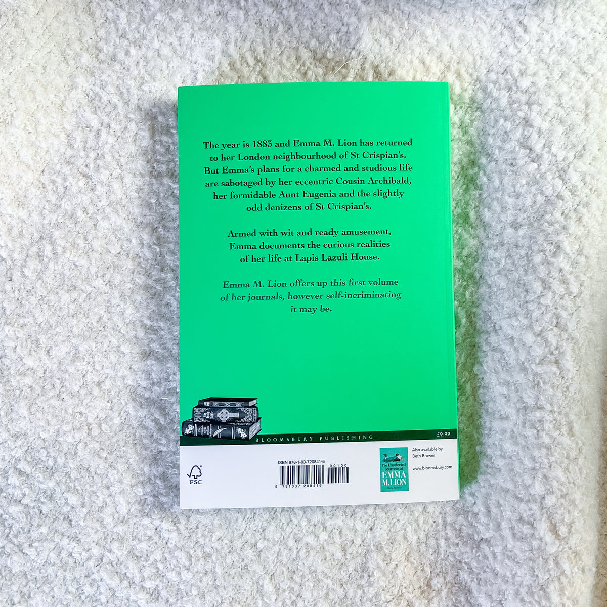 The back cover of the book 'Emma' by Emma M. Lion, featuring a bright green background with white text, a small black-and-white illustration of a horse-drawn carriage, and publisher logos at the bottom, photographed on a textured gray fabric surface.