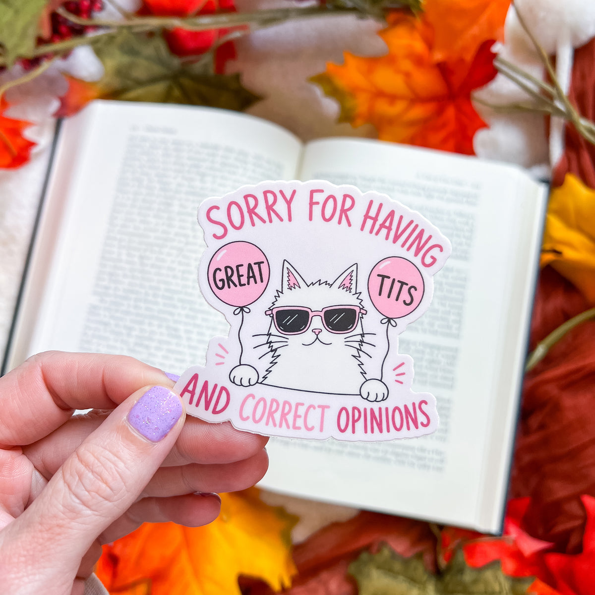 A hand with lavender glitter nail polish holds a die-cut sticker featuring a cartoon cat wearing sunglasses and holding pink balloons that say 'GREAT TITS' and 'SORRY FOR HAVING GREAT TITS AND CORRECT OPINIONS'. The sticker is positioned over an open book surrounded by autumn leaves.