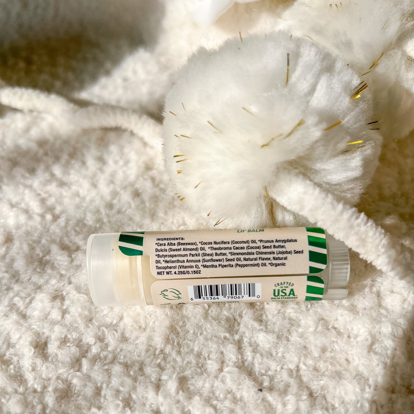 A tube of lip balm lies on a soft white textured blanket with decorative white tinsel. The label shows a white tube with green striped accents and a white cap.