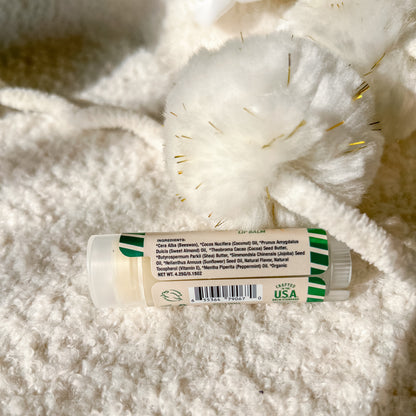A tube of lip balm lies on a soft white textured blanket with decorative white tinsel. The label shows a white tube with green striped accents and a white cap.