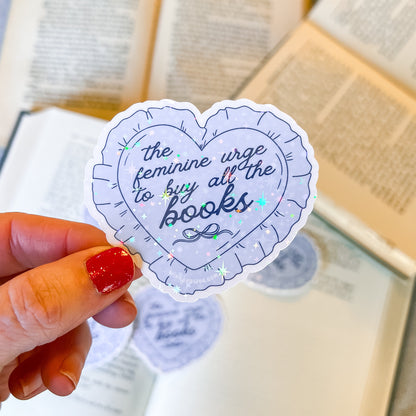 A blue heart sticker with ruffles that says “the feminine urge to bug all the books” in front of an open book.