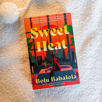 The book cover of 'Sweet Heat' by Bolu Babalola is displayed flat on a textured white blanket. The cover features a vibrant red-orange background with a city skyline silhouette, green tropical leaves, and a woman sitting on a bed.