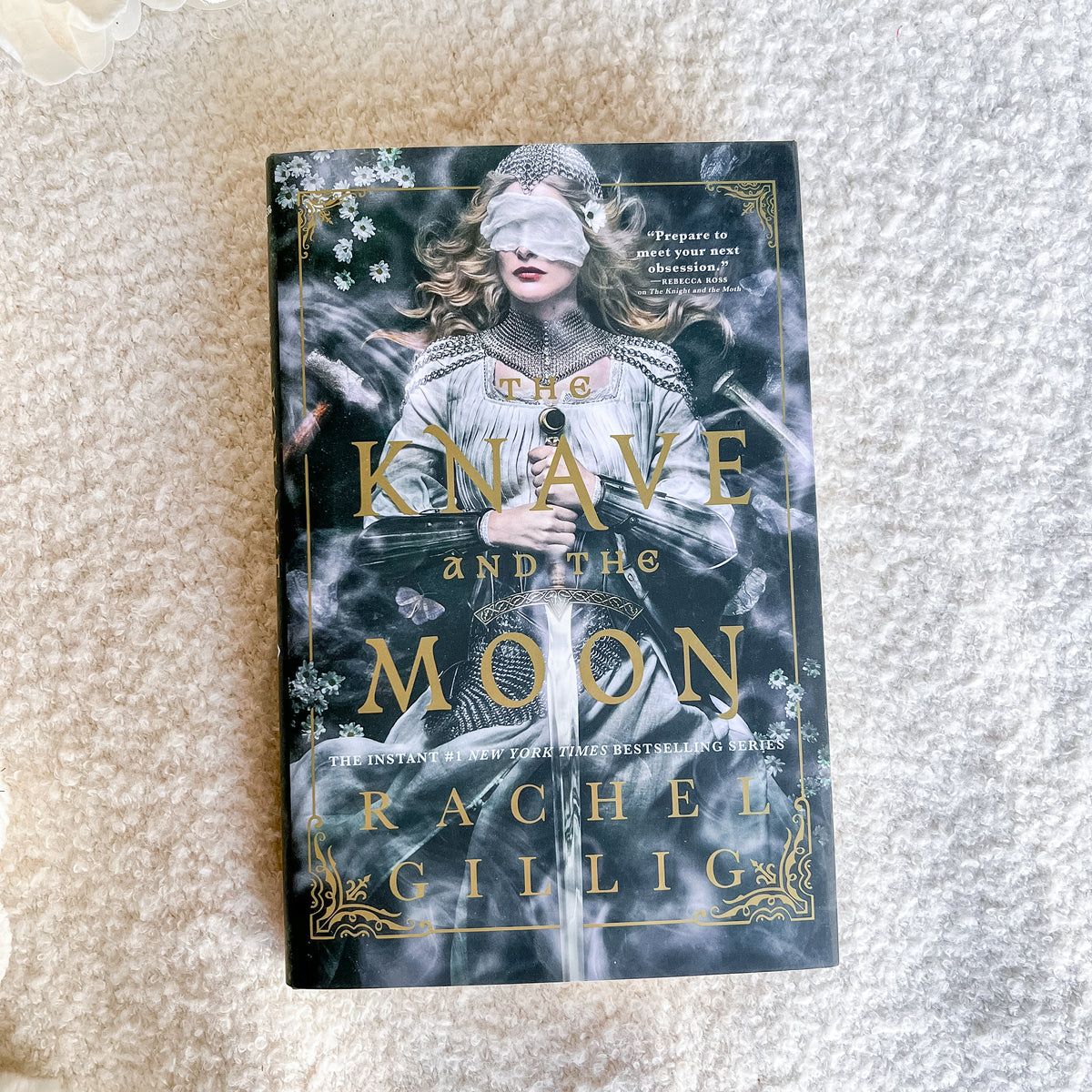 The Knave and the Moon book by Rachel Gillig, featuring a dark cover with a blindfolded girl in a white dress and gold lettering, placed on a textured cream-colored carpet.