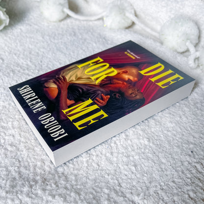 A hardcover book titled 'For Me Die' by Shiklène Obuobi, featuring a dark cover with a couple embracing and bold yellow title text, placed on a white textured blanket with decorative white pom-pom tassels in the background.