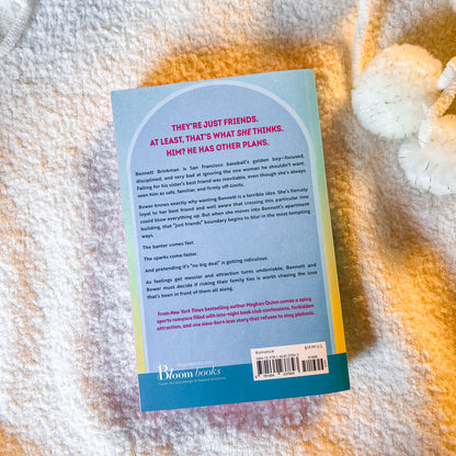 The back cover of a Bloom Books paperback novel, 'They're Just Friends,' by Rachel Ross, photographed flat on a textured cream-colored blanket.