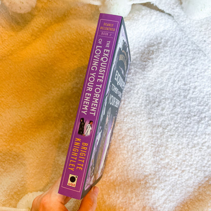 A hand holds a book titled 'The Exquisite Tortment of Loving Your Enemy' by Britette Knightley, Book 2 of the Pearls Beguiled series. The book spine is purple with gold text and white title lettering, set against a soft, textured cream and yellow blanket background.