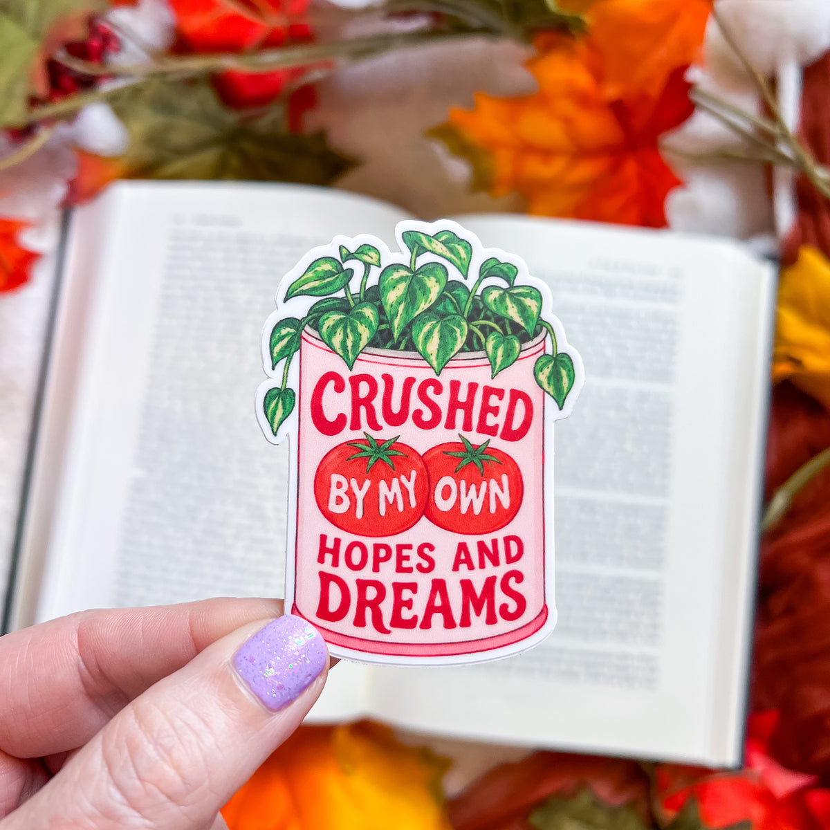 A hand with lavender glitter nail polish holds a die-cut sticker featuring a pink can with green leafy vines and red tomatoes, with the text 'CRUSHED BY MY OWN HOPES AND DREAMS'. The background shows an open book surrounded by autumn leaves.