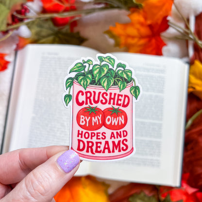 A hand with lavender glitter nail polish holds a die-cut sticker featuring a pink can with green leafy vines and red tomatoes, with the text 'CRUSHED BY MY OWN HOPES AND DREAMS'. The background shows an open book surrounded by autumn leaves.