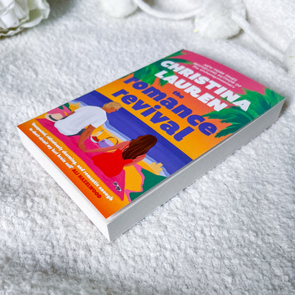 The book 'The Romance of Rival' by Christina Lauren is shown lying on a white textured blanket. The cover features a vibrant, colorful illustration with a pink sky, blue water, and a yellow-orange foreground, with the author's name in bold white letters.