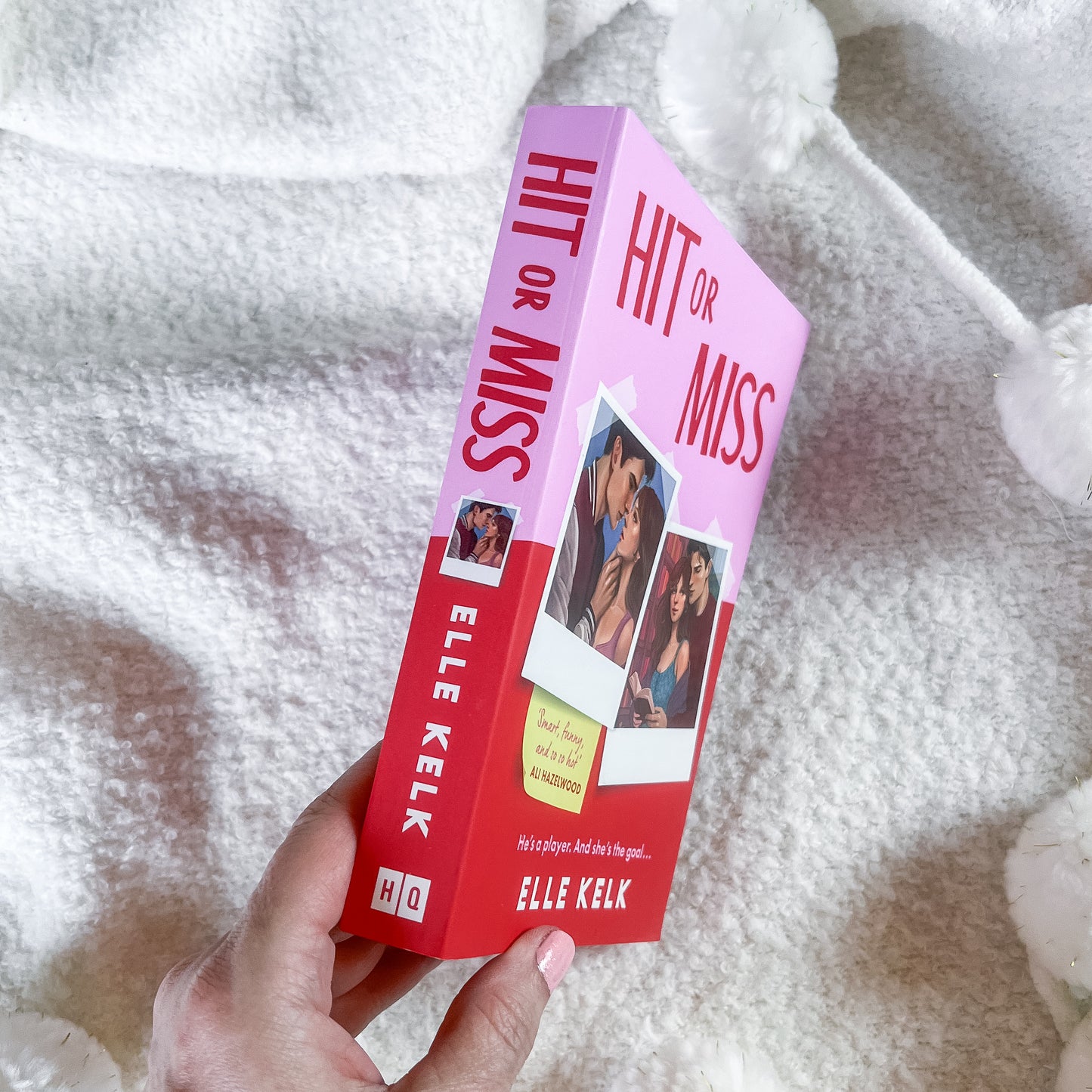 A hand holds a paperback book titled 'Hit or Miss' by Elle Kelk. The book cover is pink and red with white text and small photos of a couple, set against a soft white textured blanket background.