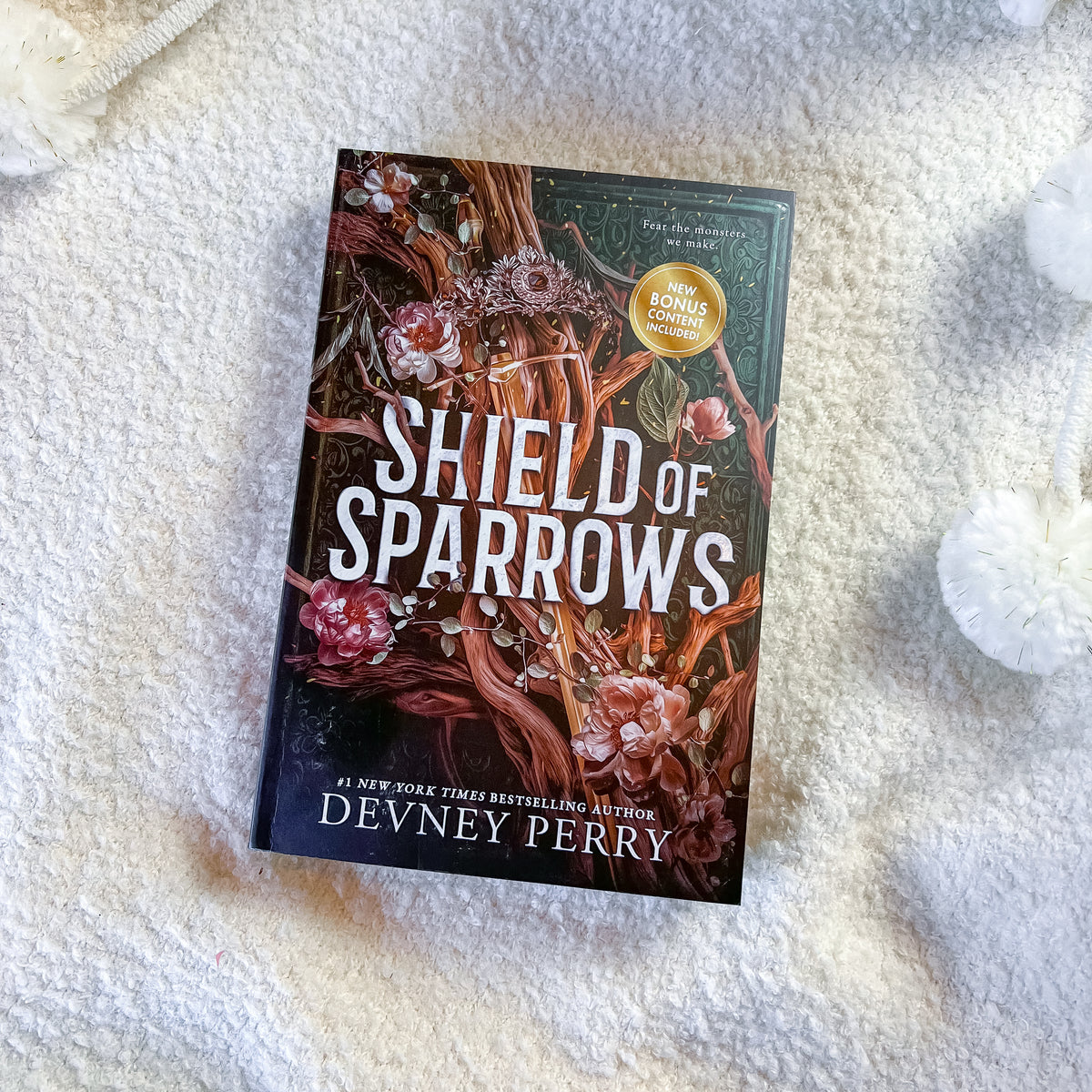 The book 'Shield of Sparrows' by Devney Perry is displayed flat on a textured white blanket, surrounded by decorative white pom-pom cords. The cover features a dark forest illustration with branches, pink flowers, and bold white title text.