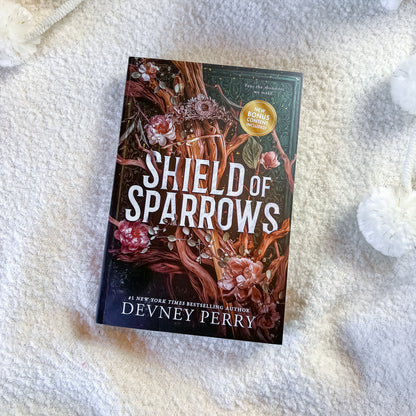 The book 'Shield of Sparrows' by Devney Perry is displayed flat on a textured white blanket, surrounded by decorative white pom-pom cords. The cover features a dark forest illustration with branches, pink flowers, and bold white title text.