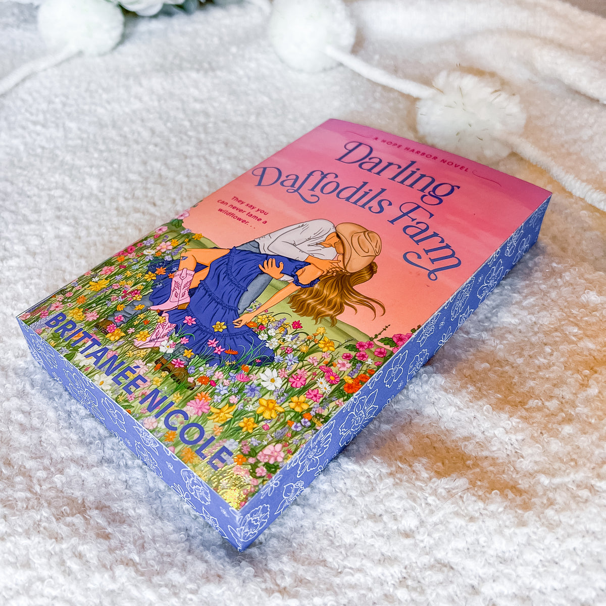 The book 'Darling Daffodils Farm' by Brittany Nicole is displayed diagonally on a soft, textured white blanket with decorative white pom-pom flowers in the background. The book cover features a colorful illustration of a girl in a blue dress among daffodils under a pink and orange sky, with a blue patterned spine.
