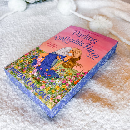 The book 'Darling Daffodils Farm' by Brittany Nicole is displayed diagonally on a soft, textured white blanket with decorative white pom-pom flowers in the background. The book cover features a colorful illustration of a girl in a blue dress among daffodils under a pink and orange sky, with a blue patterned spine.