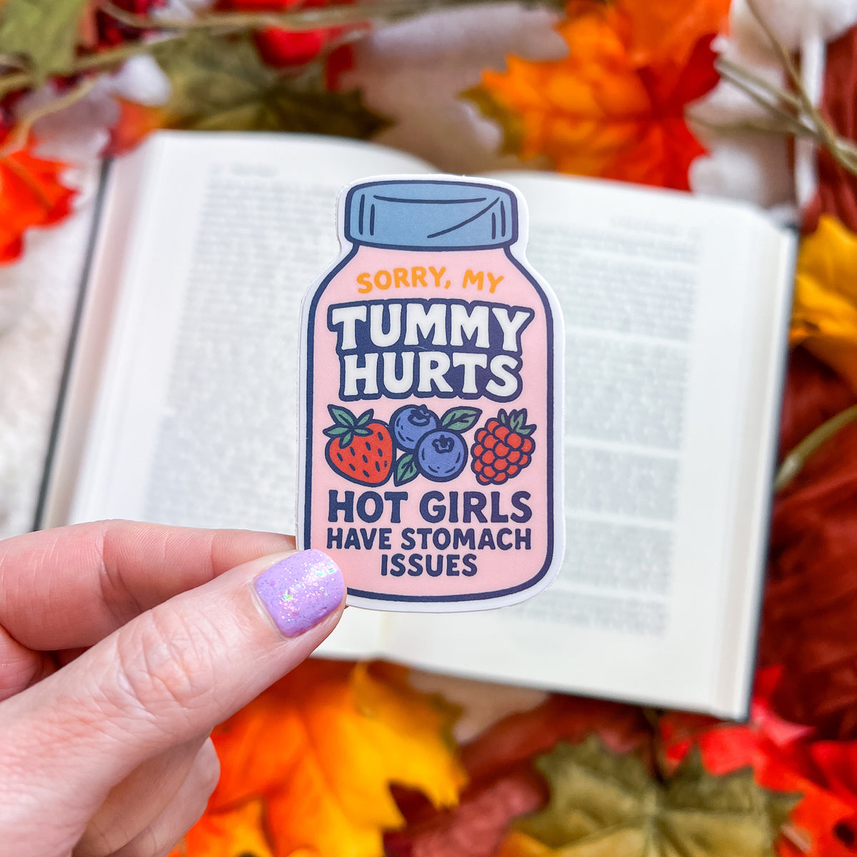 A hand with lavender glitter nail polish holds a die-cut sticker shaped like a medicine bottle. The sticker has a pink body, a blue cap, and colorful illustrations of strawberries, blueberries, and raspberries, with the text 'SORRY, MY TUMMY HURTS HOT GIRLS HAVE STOMACH ISSUES'.