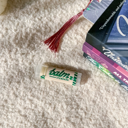 A tube of Balm Standard lip balm lies on a white textured blanket next to a stack of books and a red tassel bookmark.