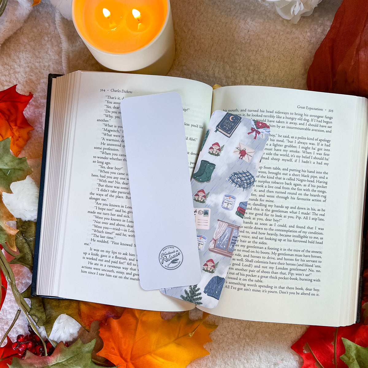 A white rectangular bookmark with a circular logo sits on an open book surrounded by autumn leaves and a lit candle. The bookmark features a vertical panel of illustrated winter scenes, including snow, a scarf, and a hat.