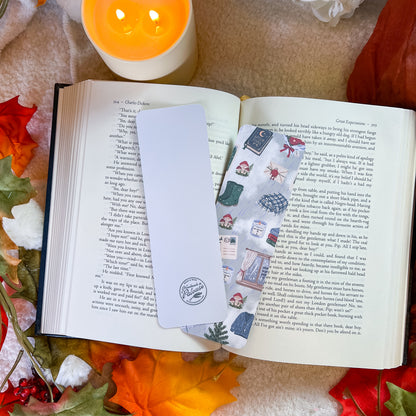 A white rectangular bookmark with a circular logo sits on an open book surrounded by autumn leaves and a lit candle. The bookmark features a vertical panel of illustrated winter scenes, including snow, a scarf, and a hat.