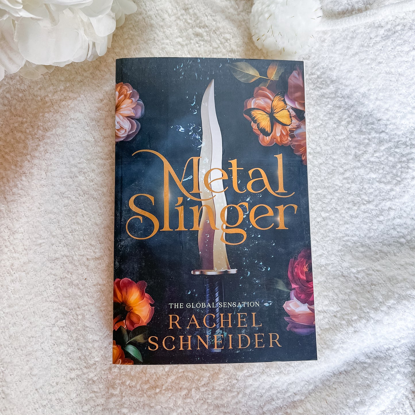 The book cover of 'Metal Slinger' by Rachel Schneider, featuring a dark background with a dagger and orange flowers, photographed on a white textured blanket.