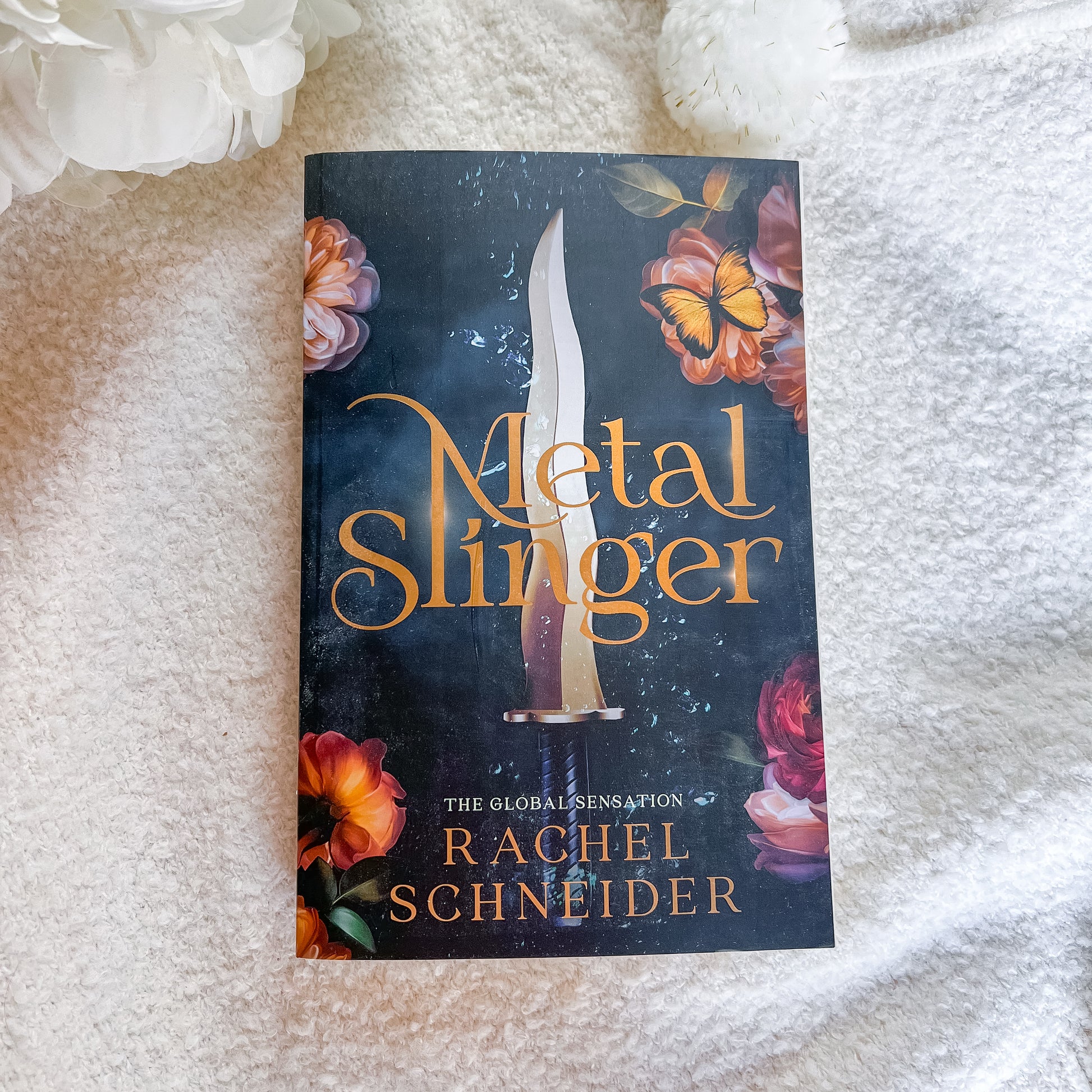 The book cover of 'Metal Slinger' by Rachel Schneider, featuring a dark background with a dagger and orange flowers, photographed on a white textured blanket.