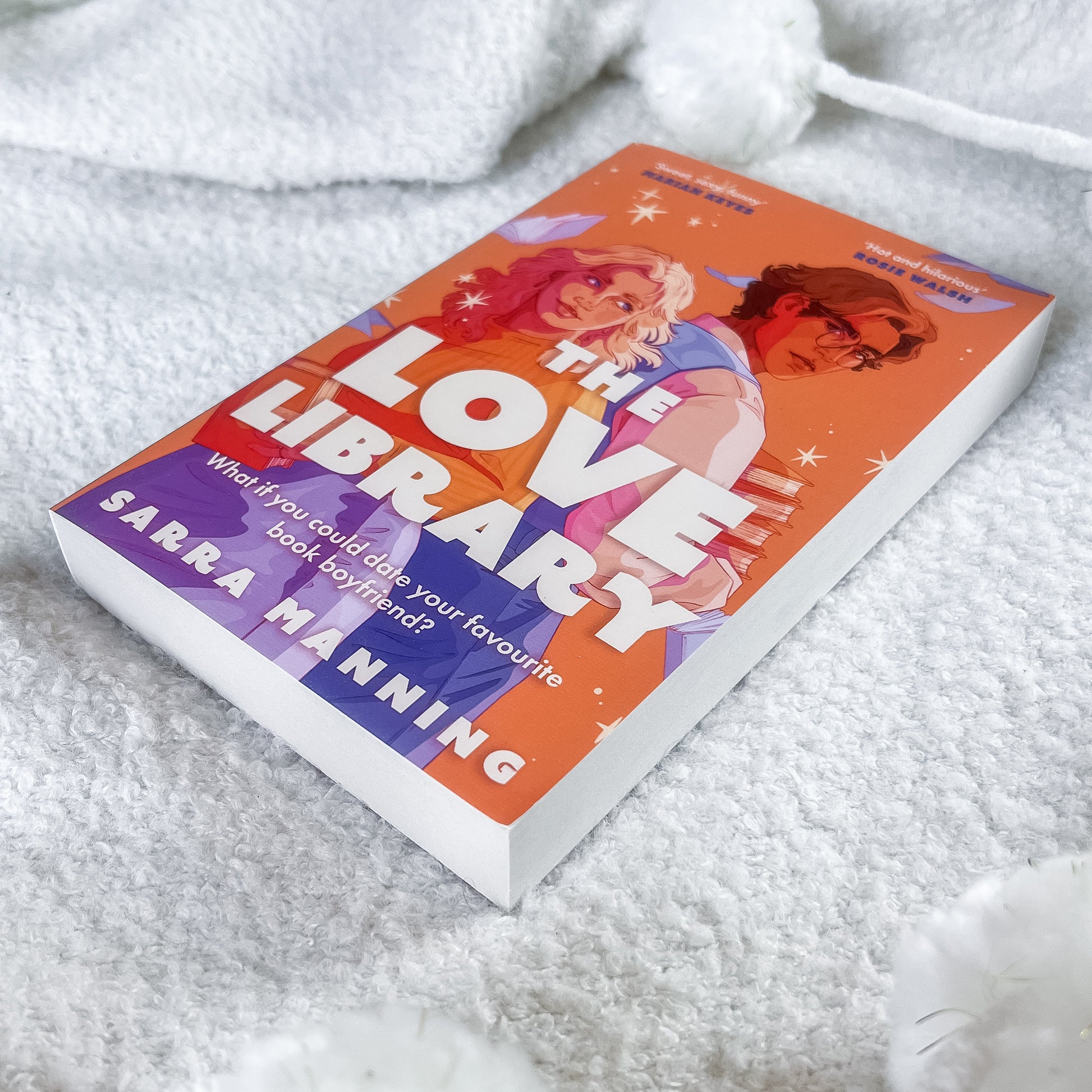 A paperback book titled "The Love Library" by Sarra Manning lies on a soft white textured blanket. The cover features a bright orange background with illustrations of two young people and a purple section at the bottom left.